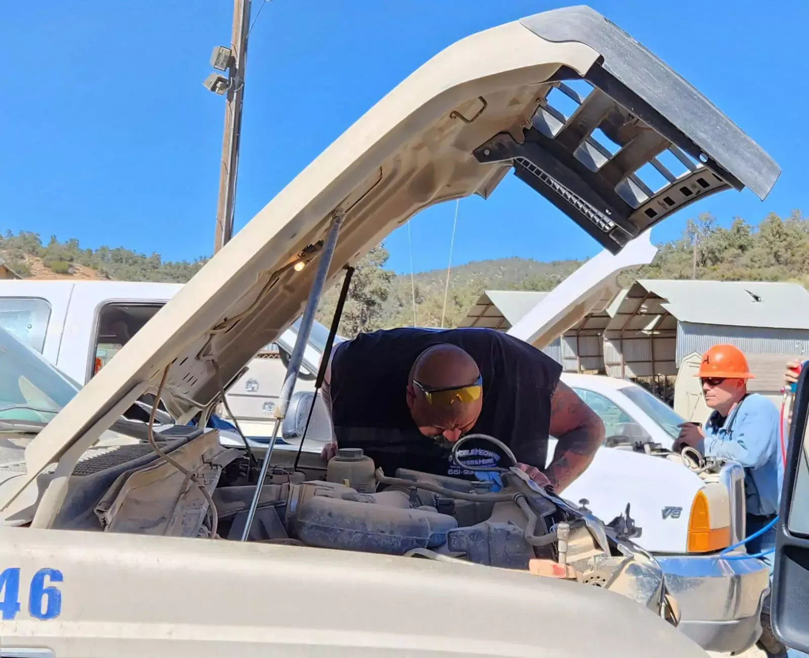 Two men working on a beige truck with the hood open outdoors on a sunny day.
