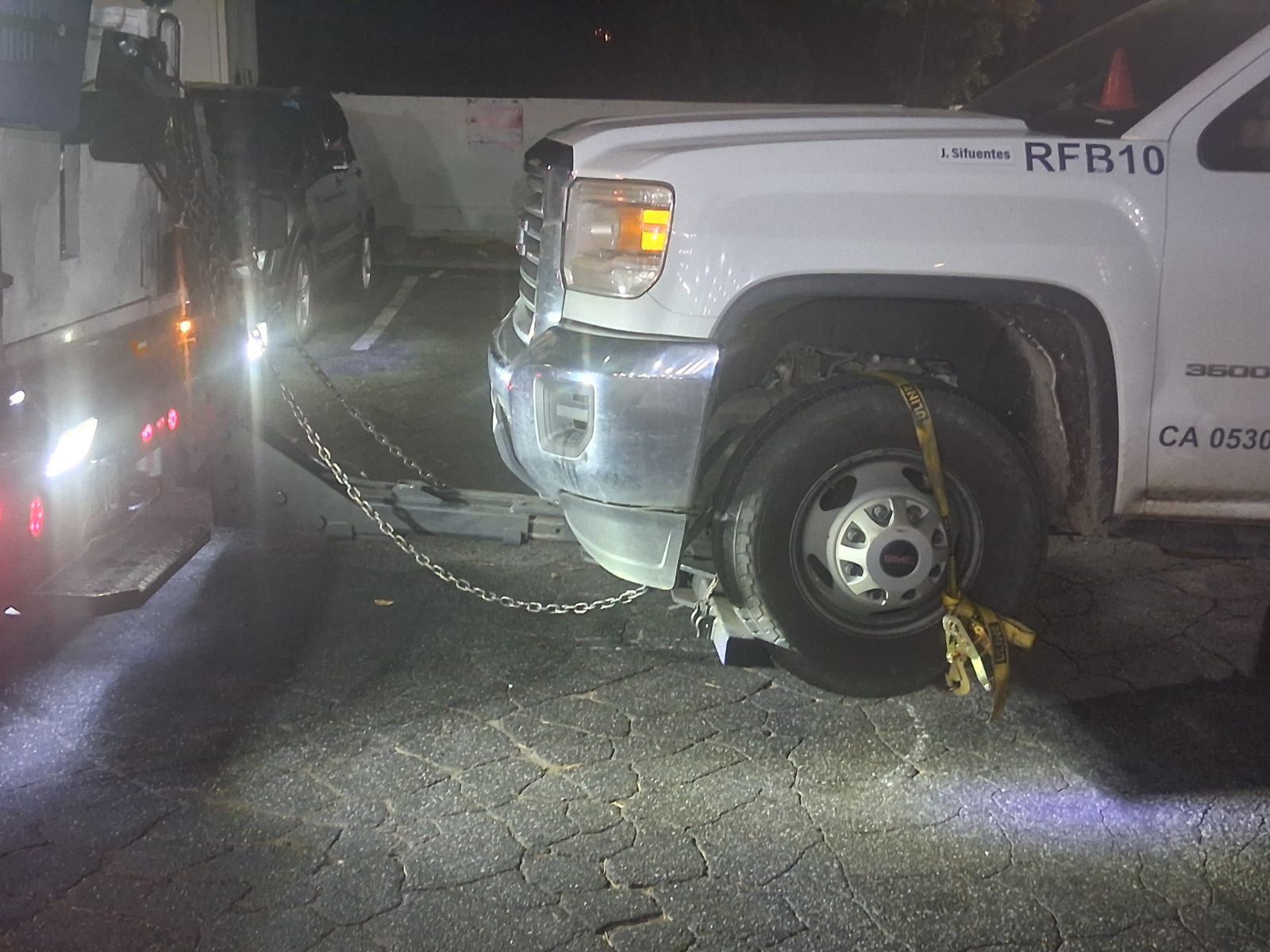 A white truck being towed by a tow truck at night. Chains and straps secure the truck's front wheel.