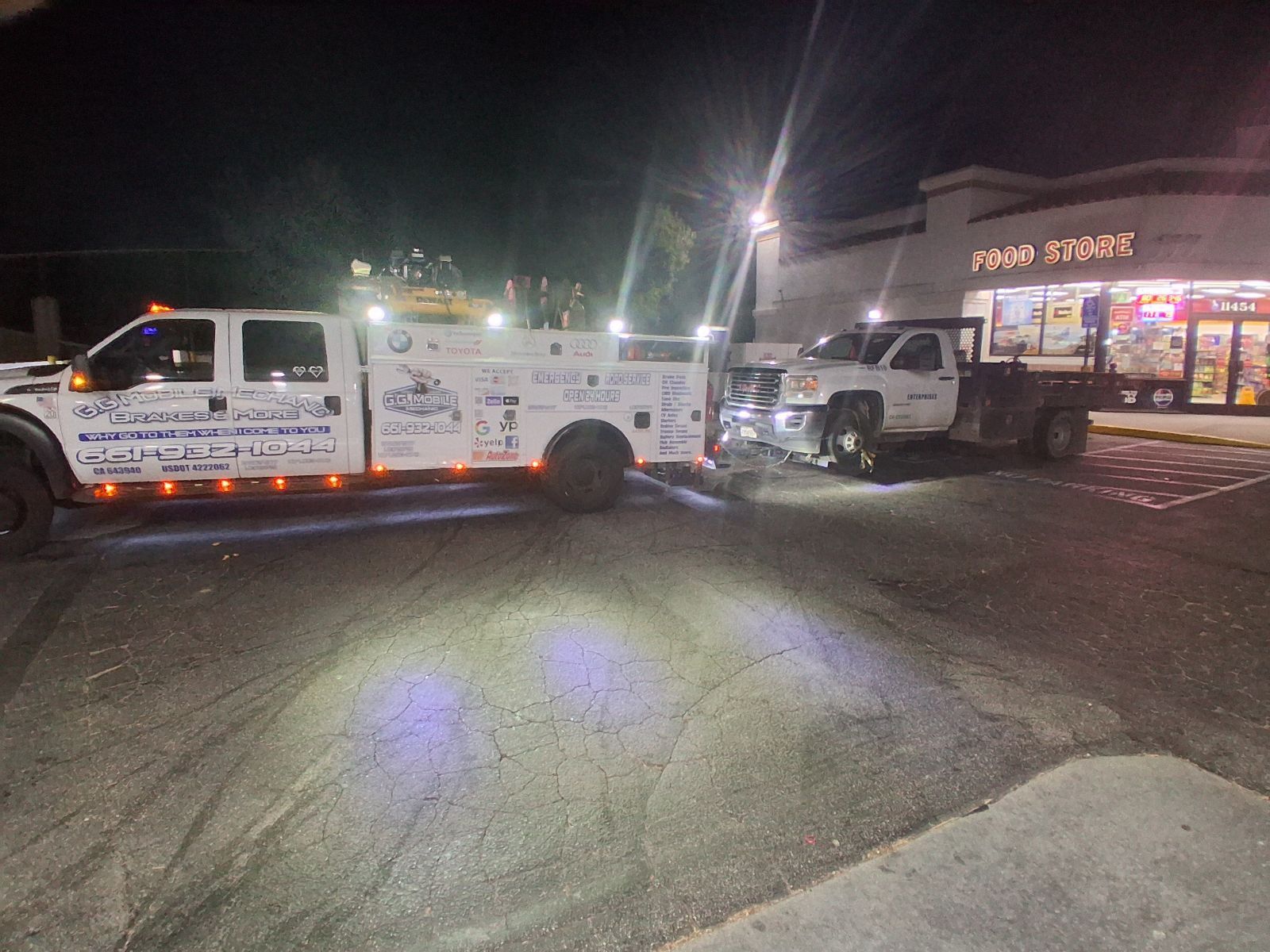 Tow truck towing a flatbed truck at night in front of a store.
