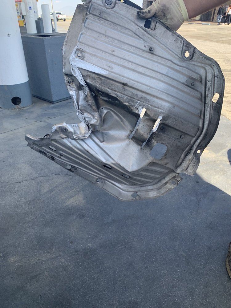 A crumpled metal car part is held by gloved hands outside a gas station.