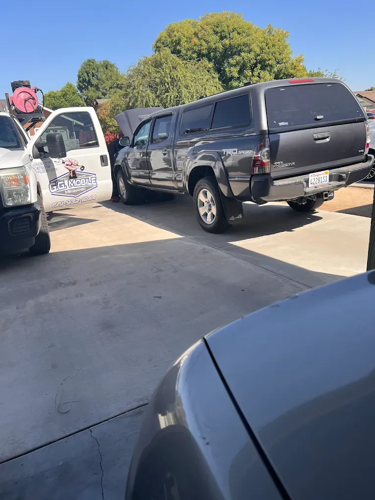 A gray Toyota pickup truck with a camper shell parked outside, hood open, next to a white service van.