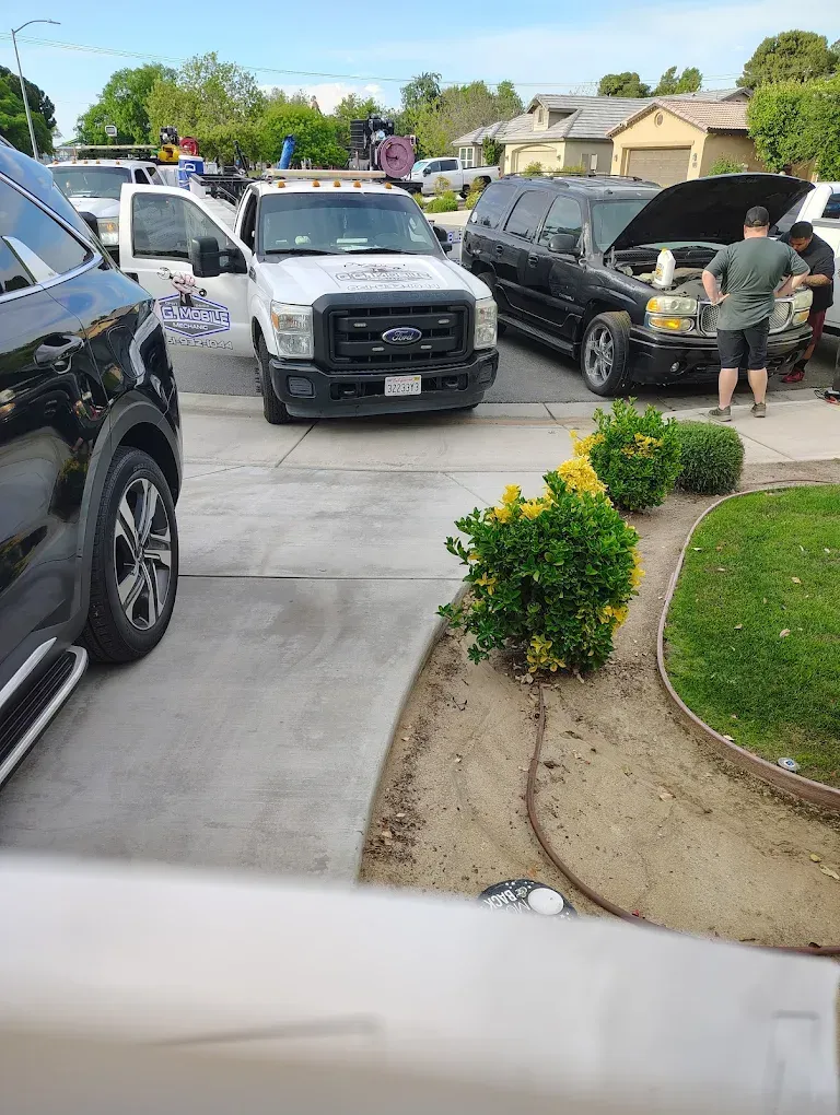 A tow truck and several vehicles are parked on a driveway. People stand near a black SUV with an open hood.