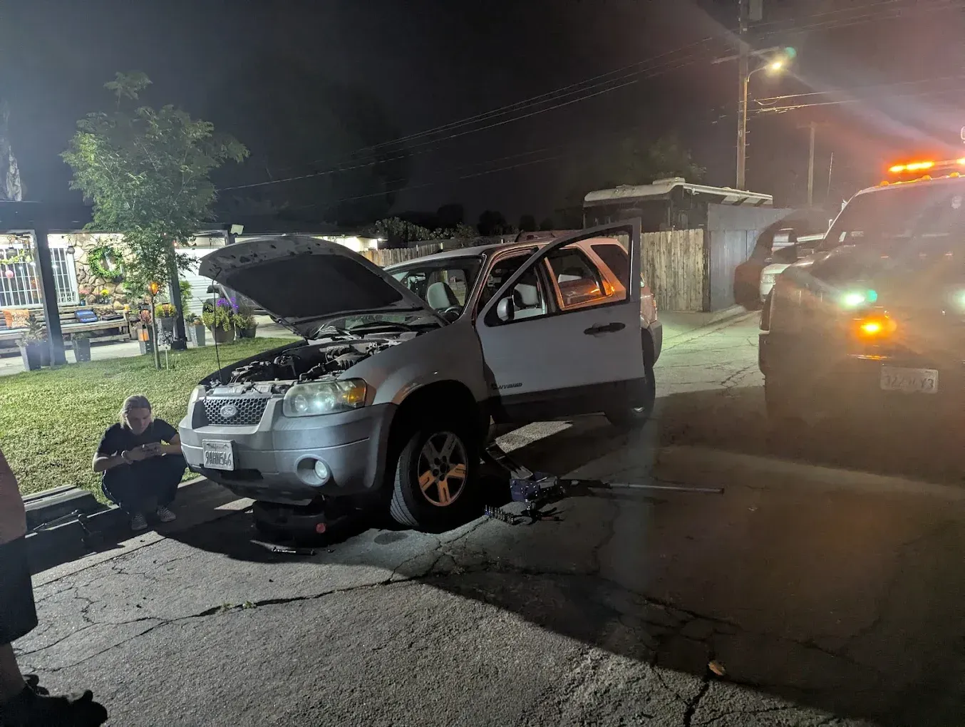 A white SUV with an open hood is on a street at night. A person is squatting nearby, and a police car is present.