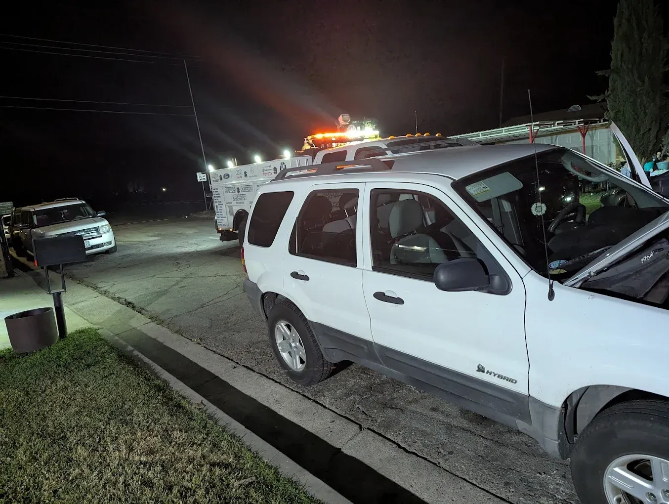 A white SUV with a damaged front end is parked near emergency vehicles at night.