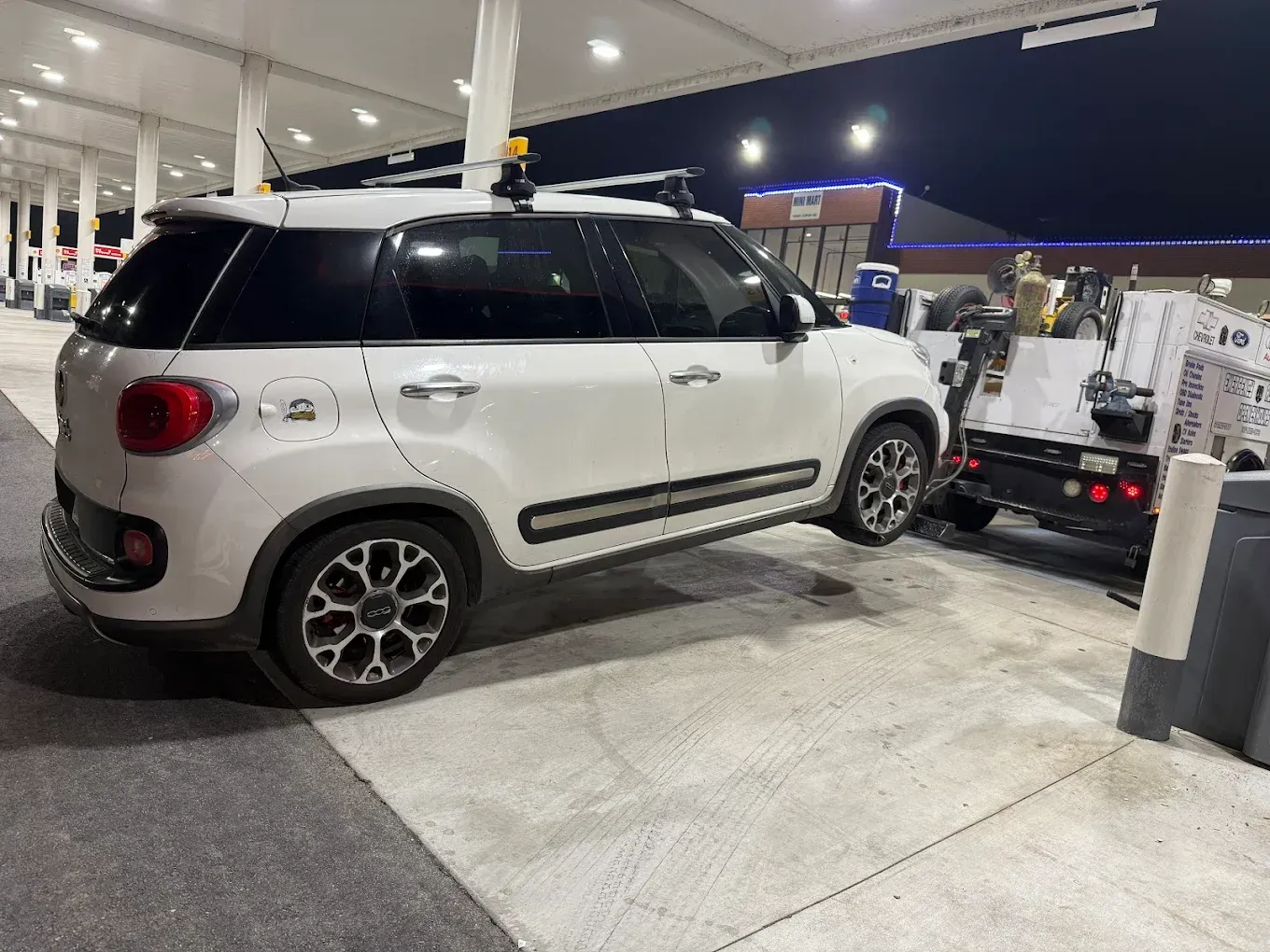 White SUV at a gas station, towing a trailer with supplies, under bright lights at night.