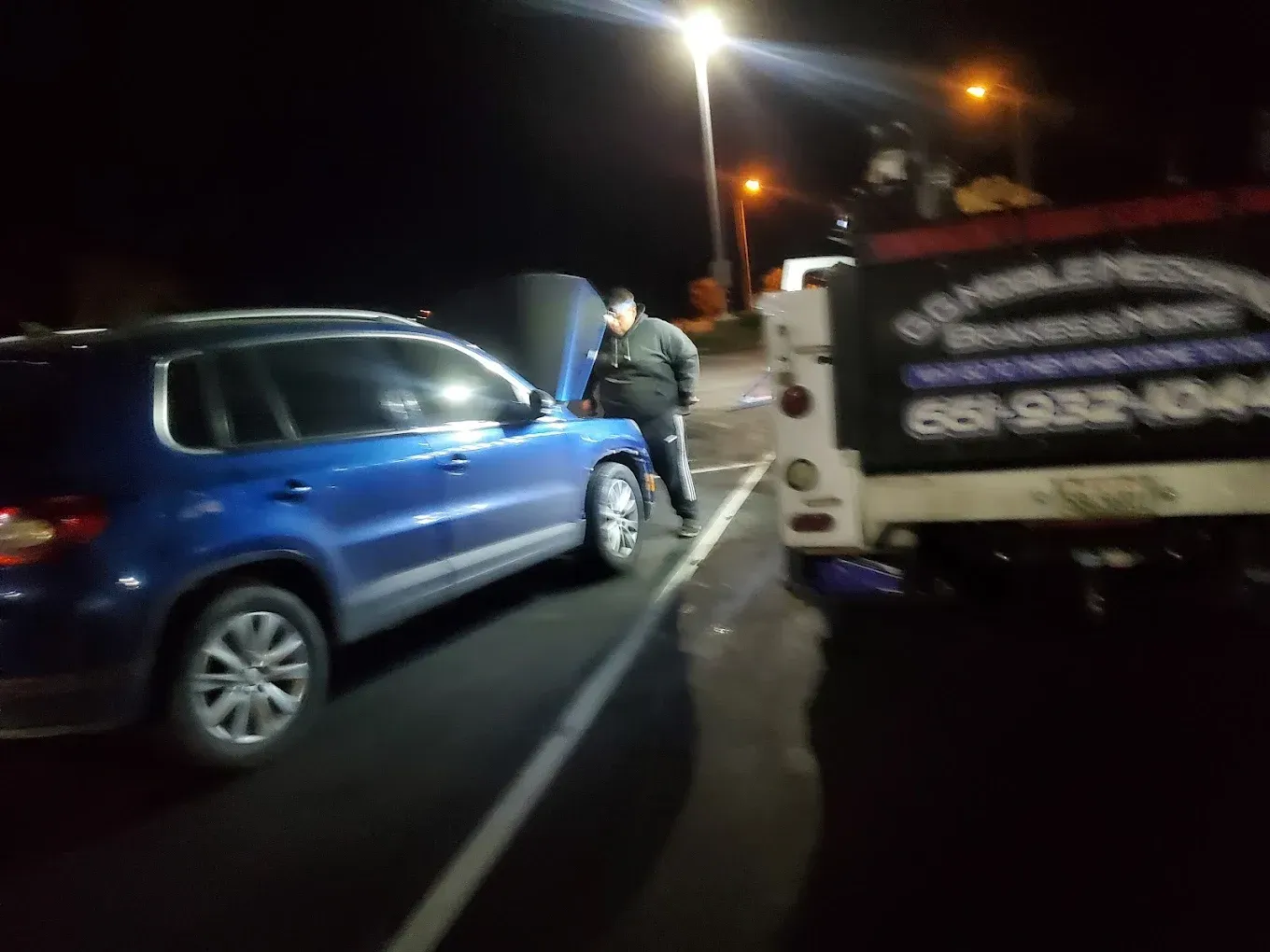 Person looking at car with an open hood next to a tow truck at night.
