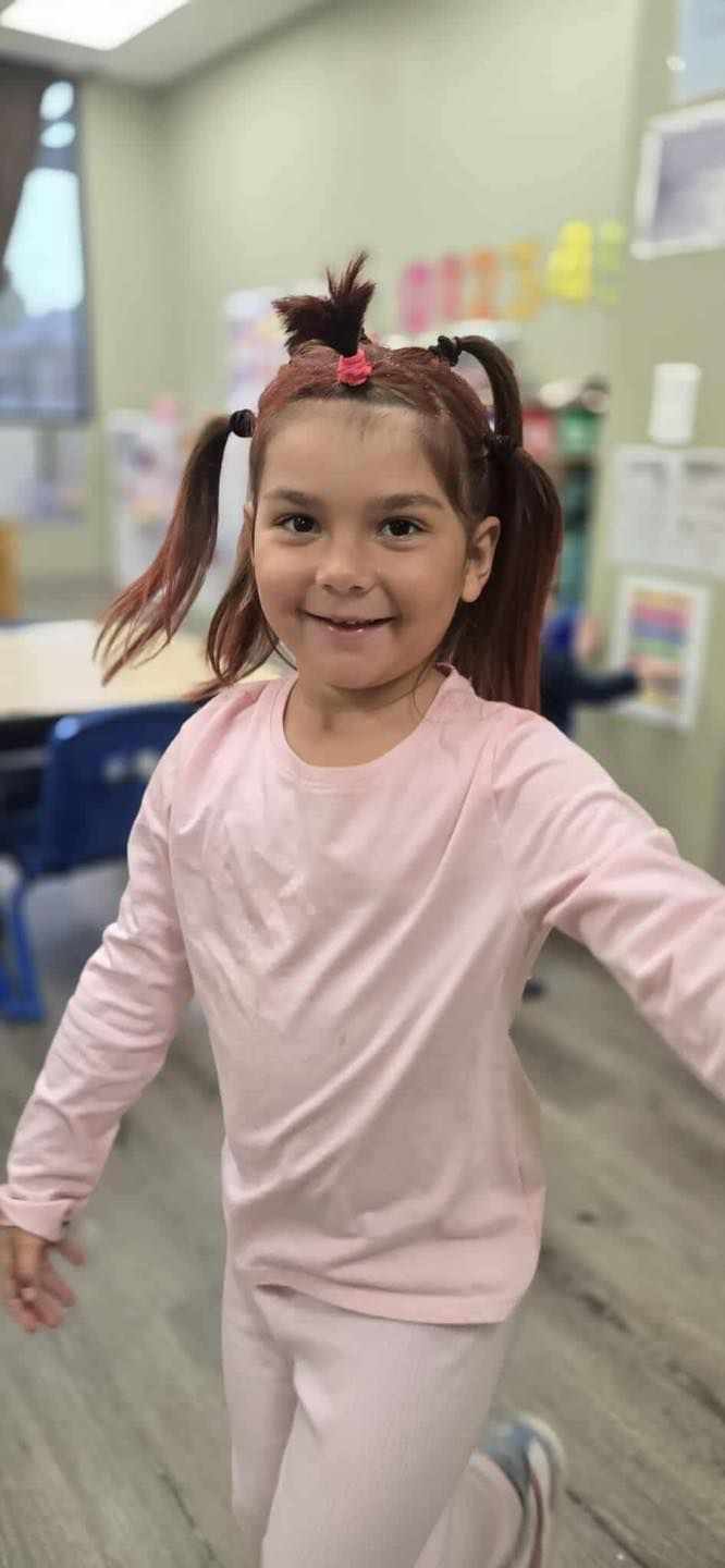 A young child wearing a light pink outfit with pigtails, smiling and posing in a room.