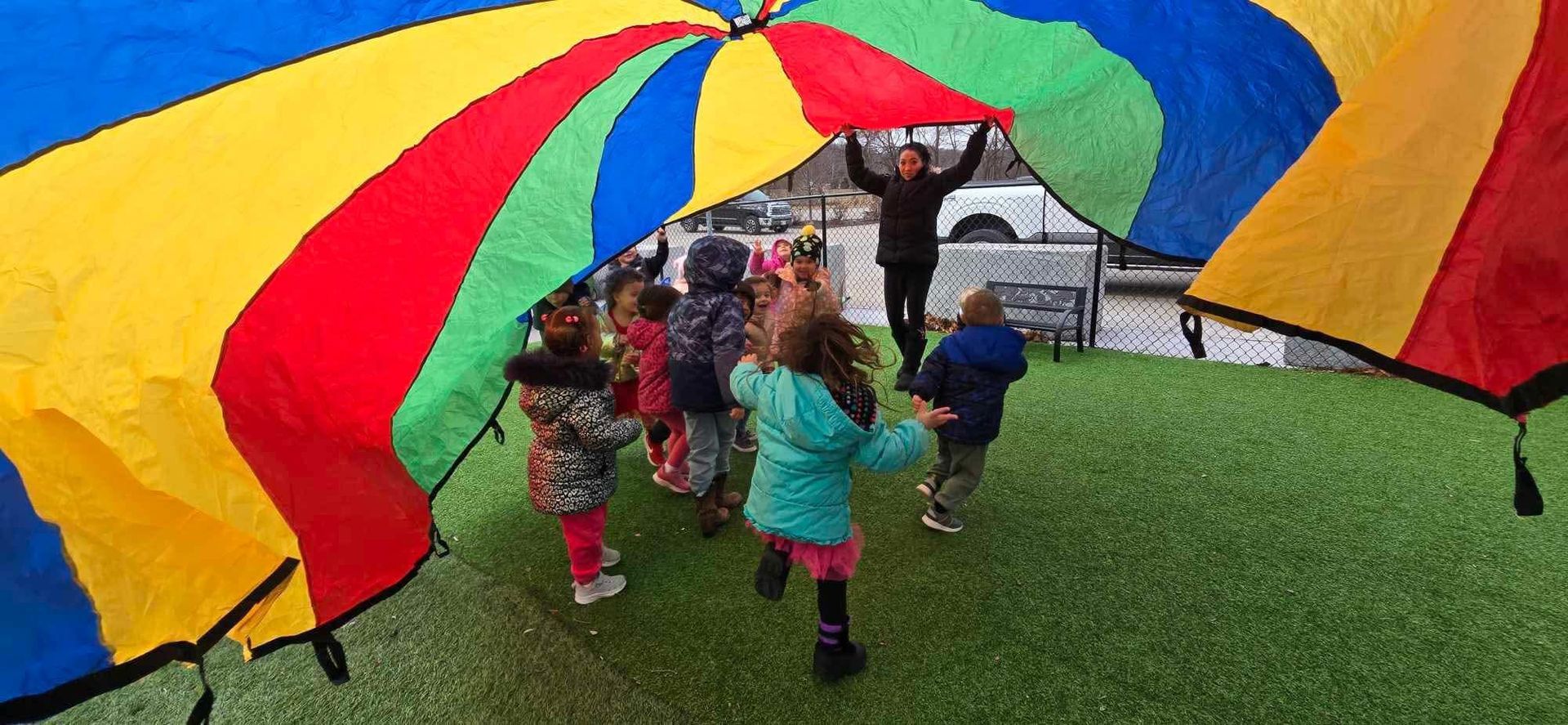 Children playing under a rainbow-colored parachute on a grassy area, with an adult supervising.