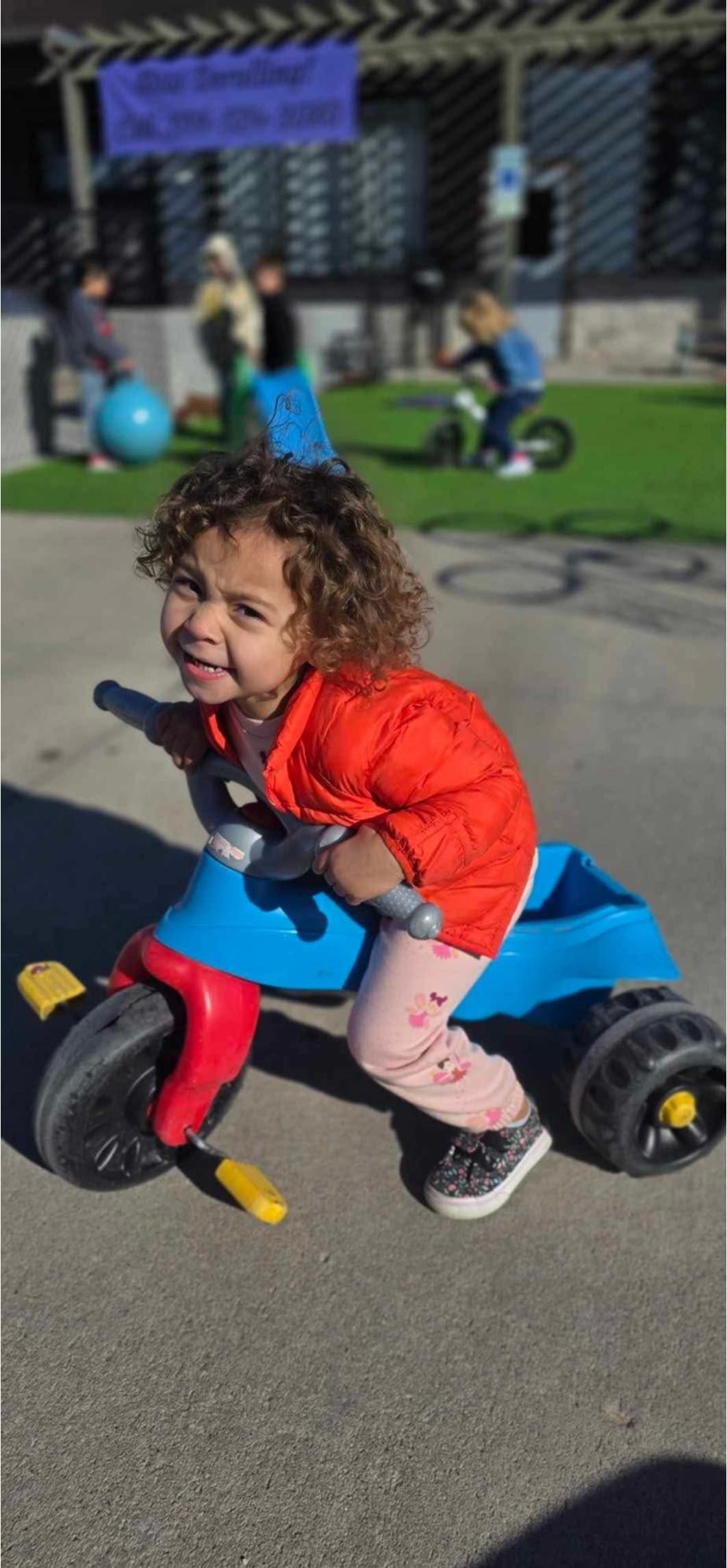 Child on a blue, red, and yellow tricycle, smiling. Outdoors, near a building.