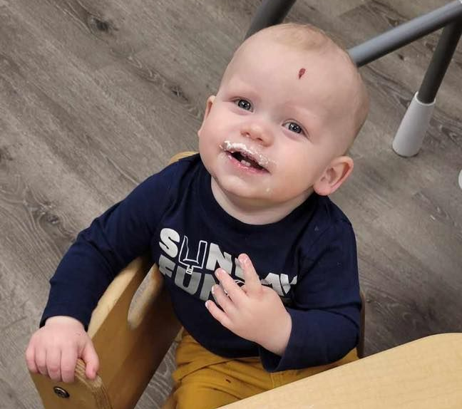 Smiling baby with food on face and shirt, sitting in a wooden highchair, wearing a blue shirt and yellow pants.