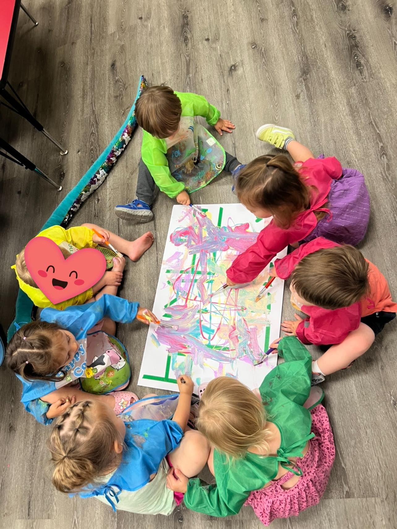 Children in a circle painting a large paper; colorful clothes, aprons, smiling.