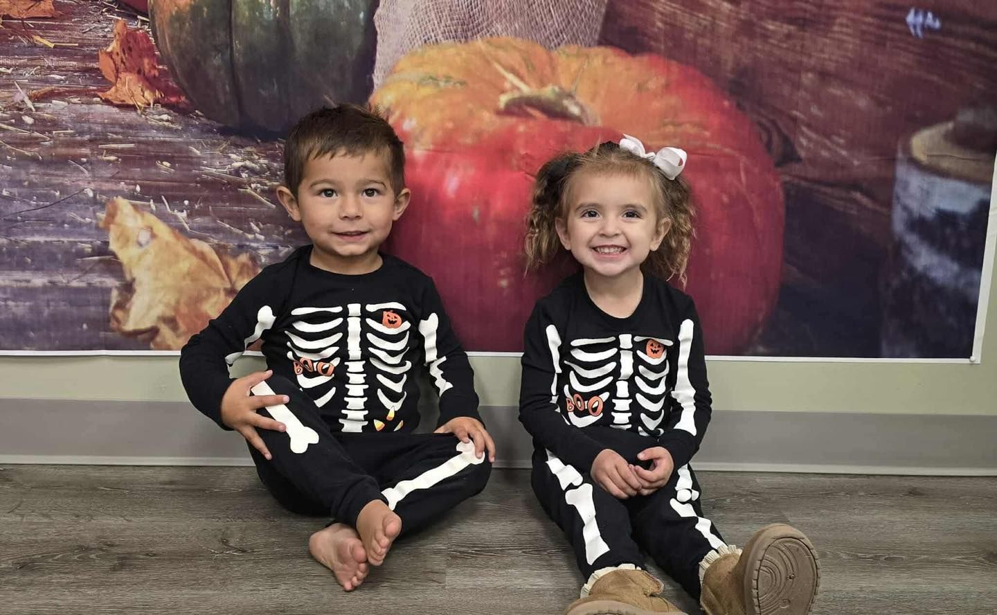 Two children in skeleton costumes sit smiling in front of a fall-themed backdrop.