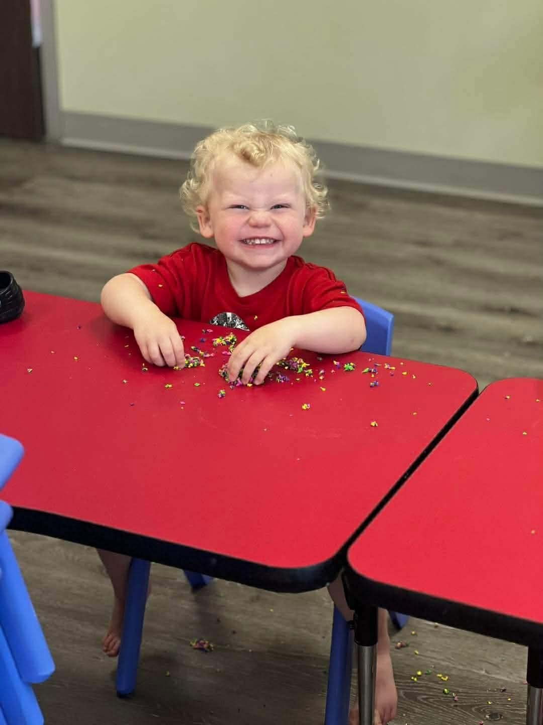 Child with curly blonde hair smiles widely at a red table, hands in scattered colorful sprinkles.