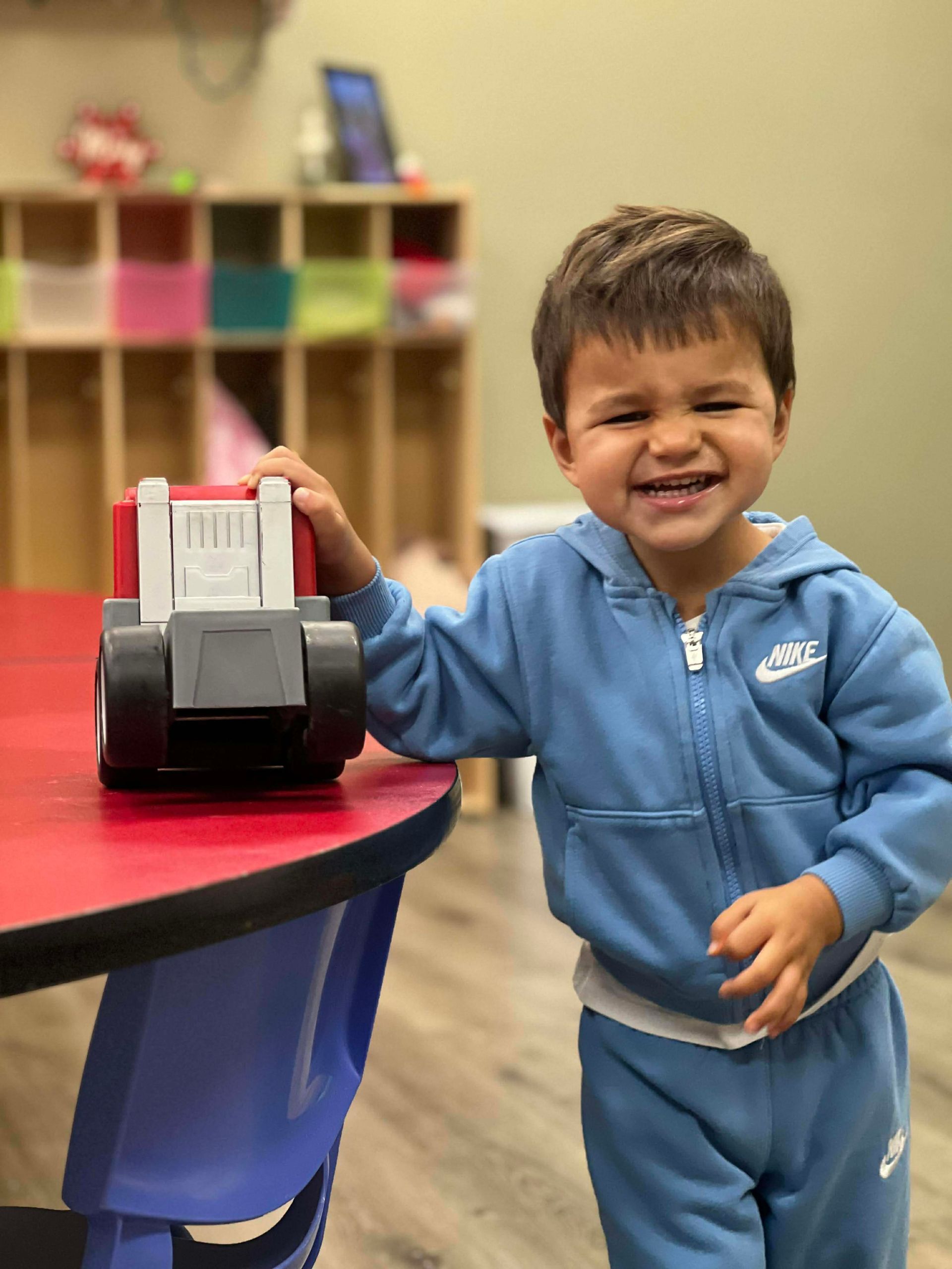 Child in blue sweatshirt smiles, holding a toy truck at a table.