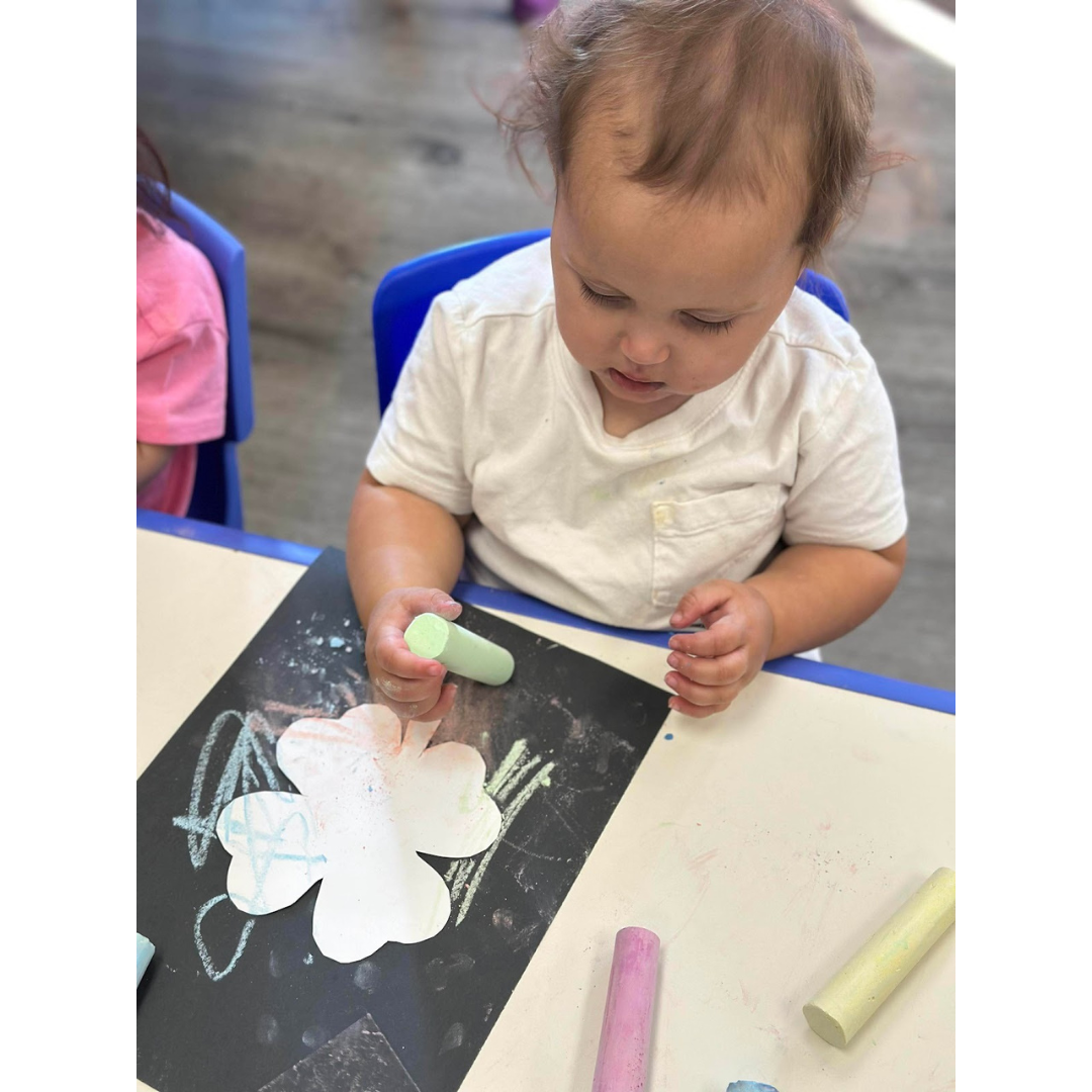 Child sitting at a table coloring a shamrock with chalk.