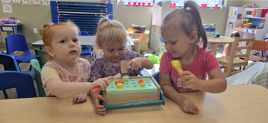 Three children play with a wooden toy at a table in a classroom.