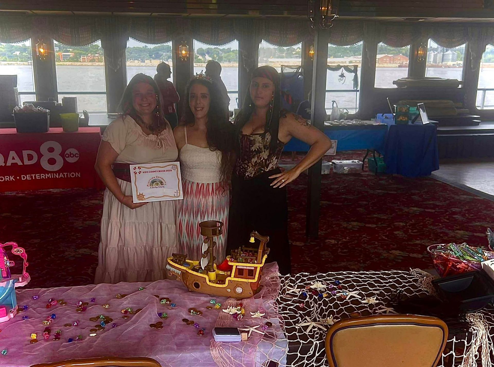 Three women pose with a pirate ship cake at an event with river views.