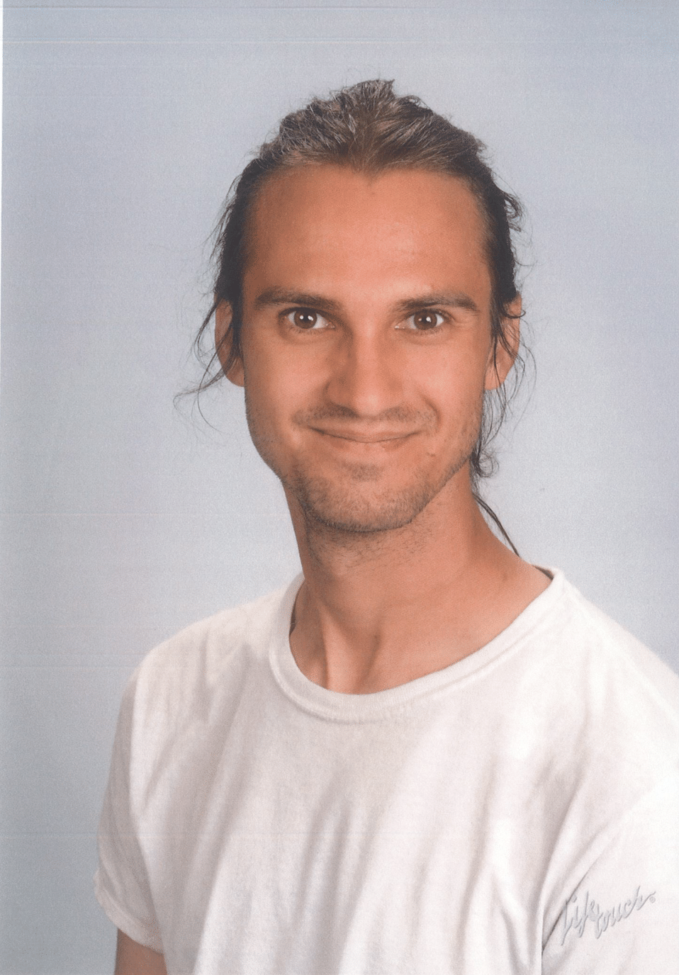 Man with long hair pulled back smiles, wearing white shirt, against a light blue backdrop.