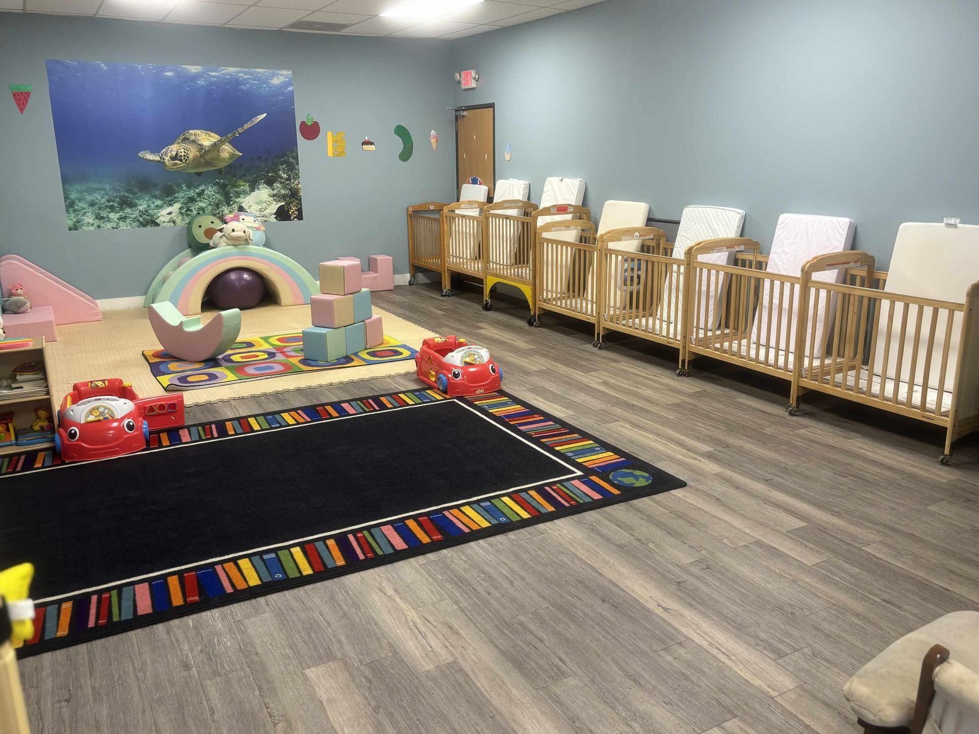 Nursery room with cribs along a wall, play area with colorful toys, and a mural of a sea turtle.
