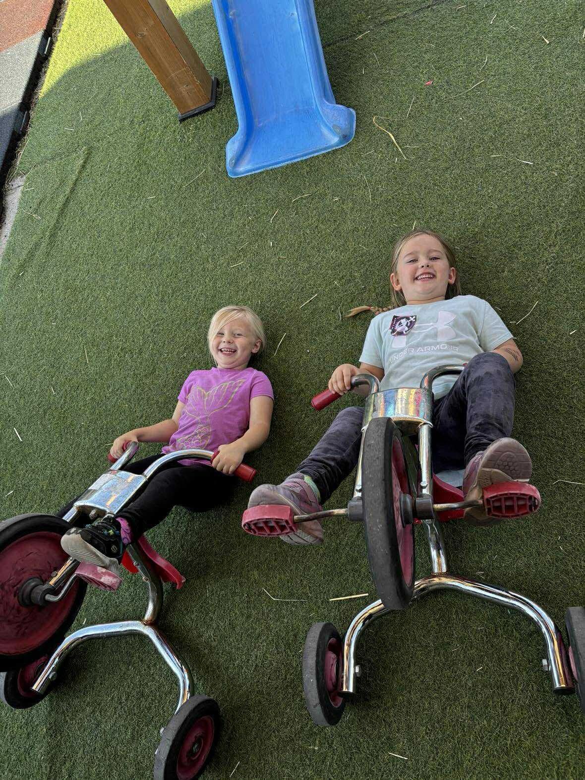 Two young girls happily riding red and silver tricycles on green turf near a blue slide.