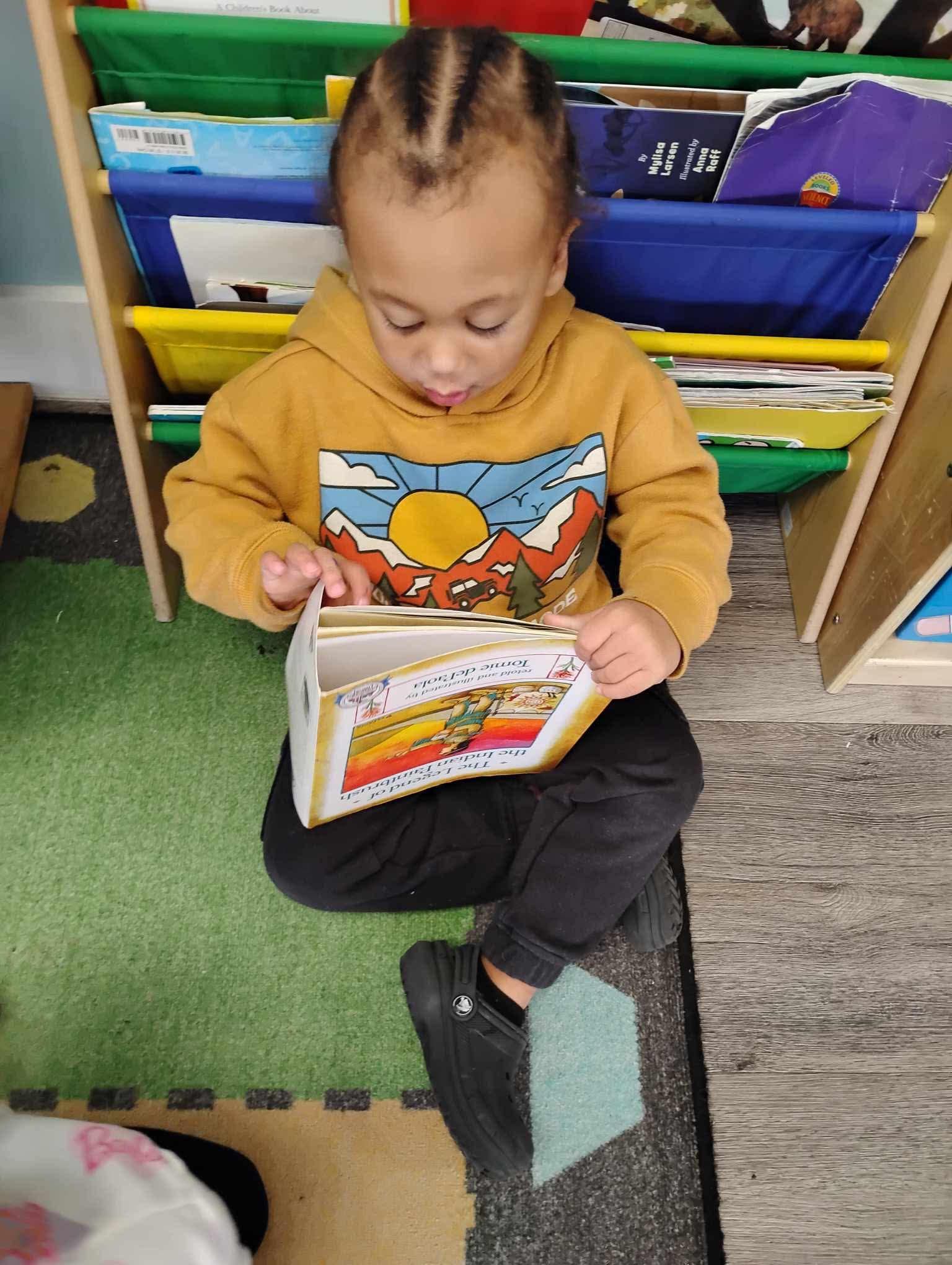 Child in yellow hoodie reading a book, sitting on a rug near bookshelves.
