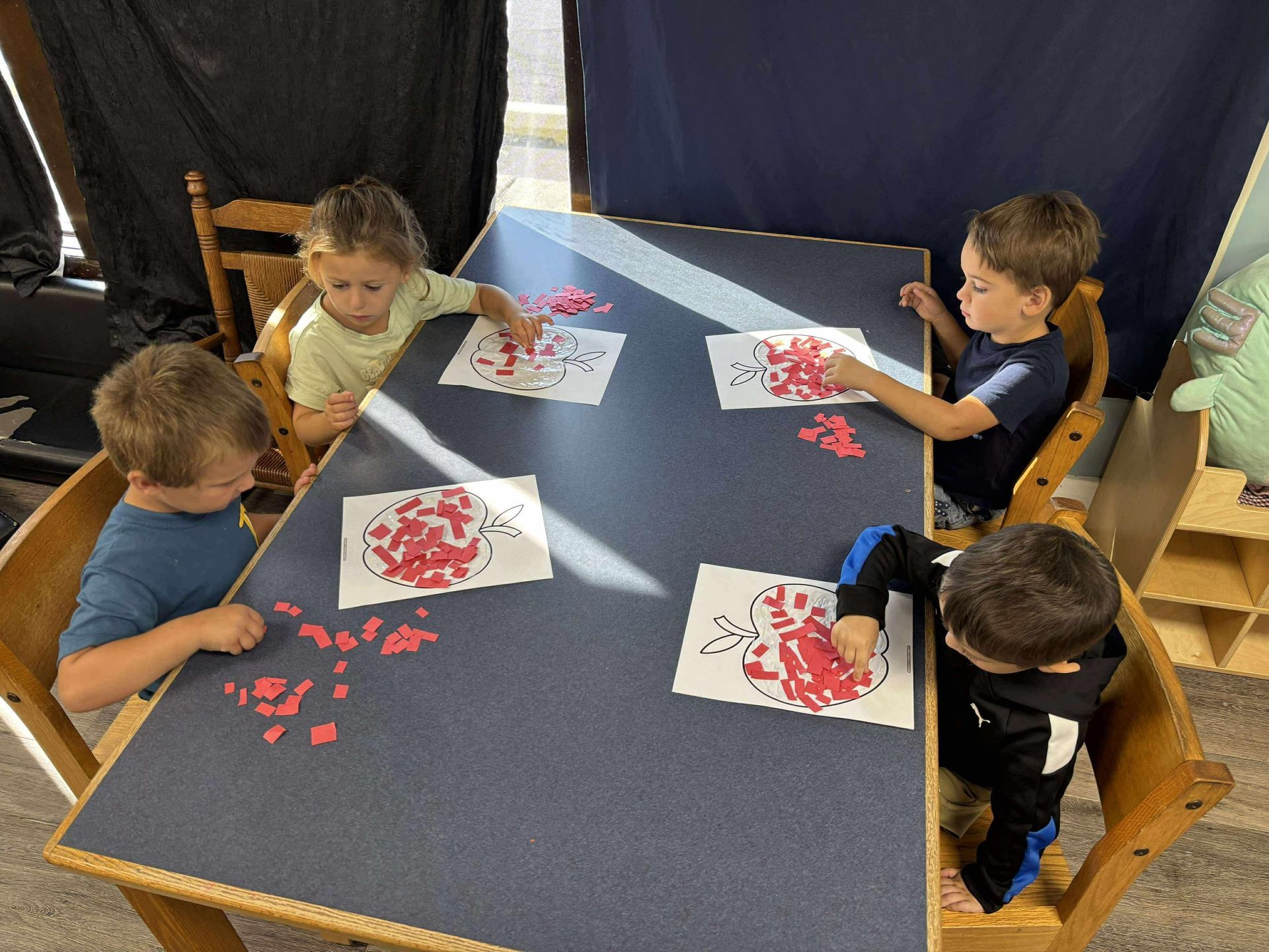 Four children at a table, gluing red shapes onto apple outlines.