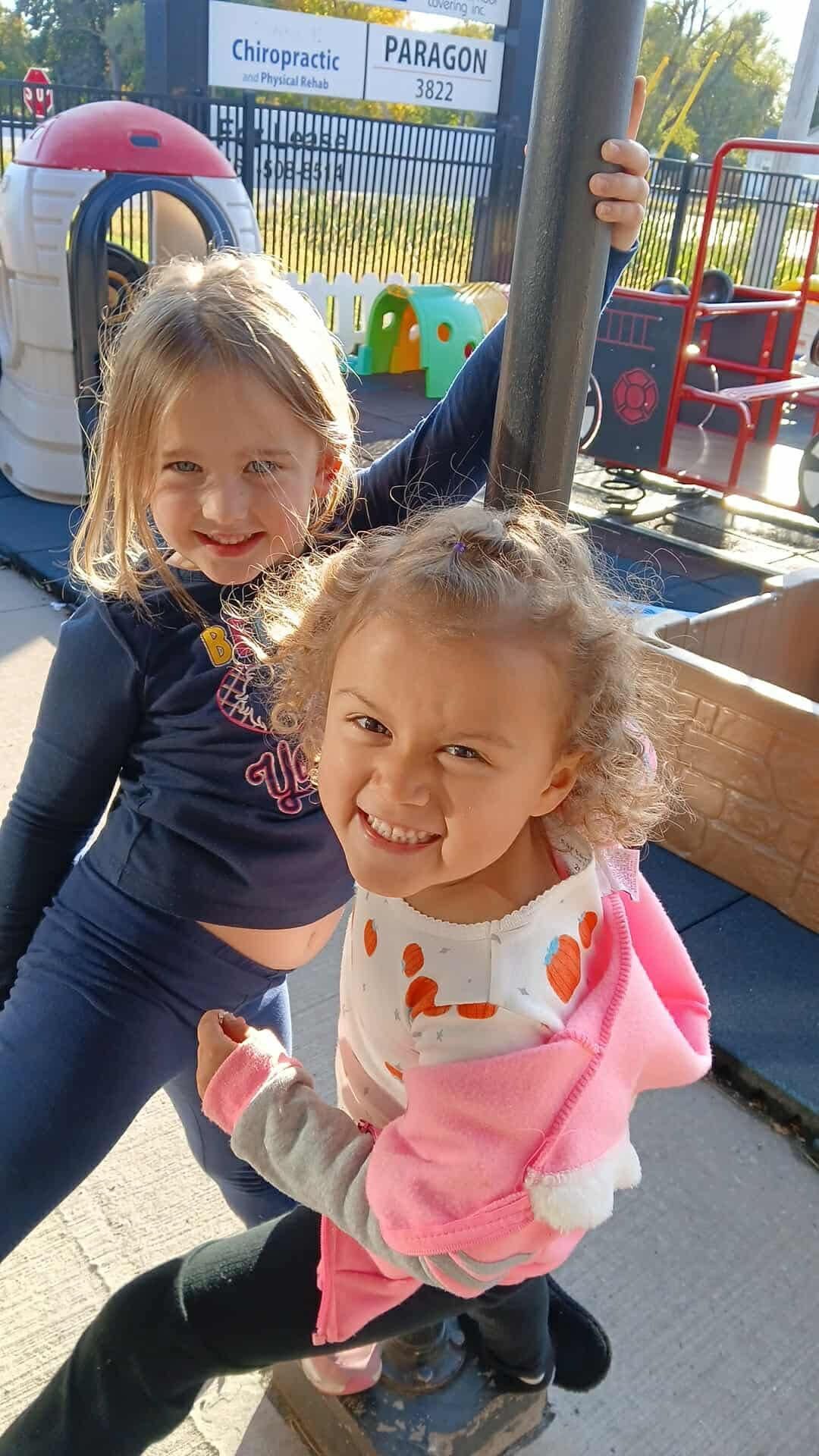 Two children smiling on playground, one holding a pole.