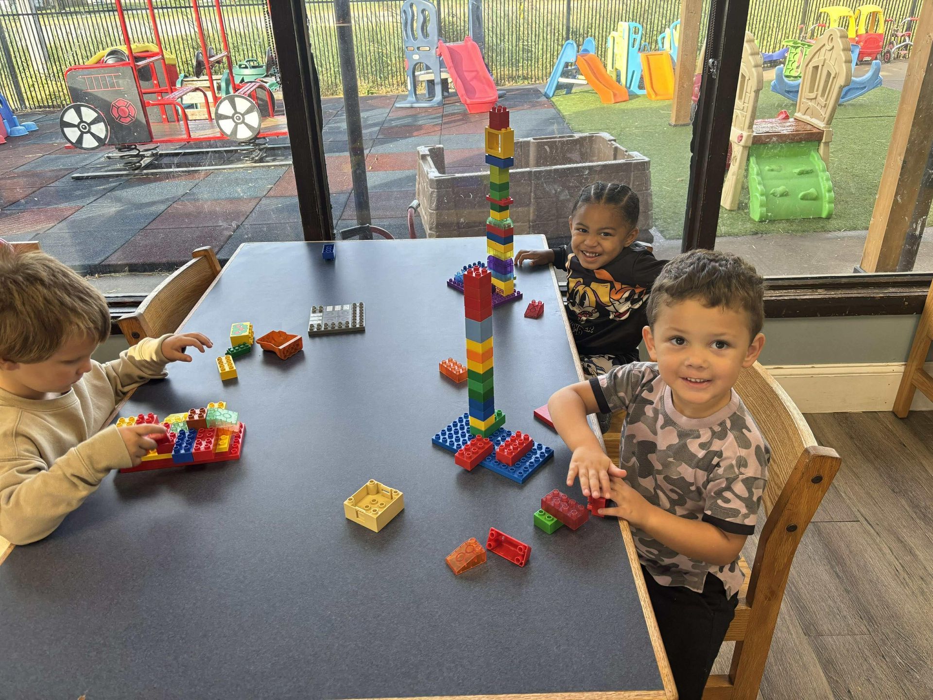 Three children smiling, playing with colorful building blocks at a table indoors, playground in background.