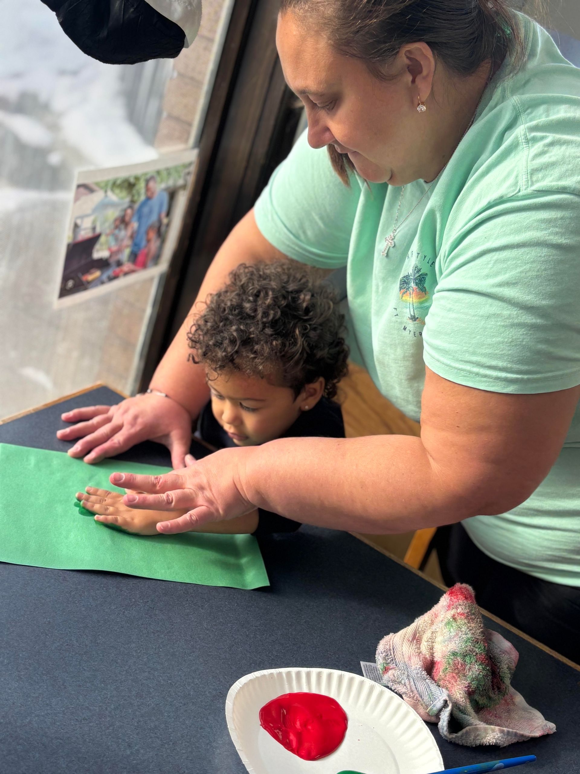 Adult assists a child with hand painting on green paper at a table.