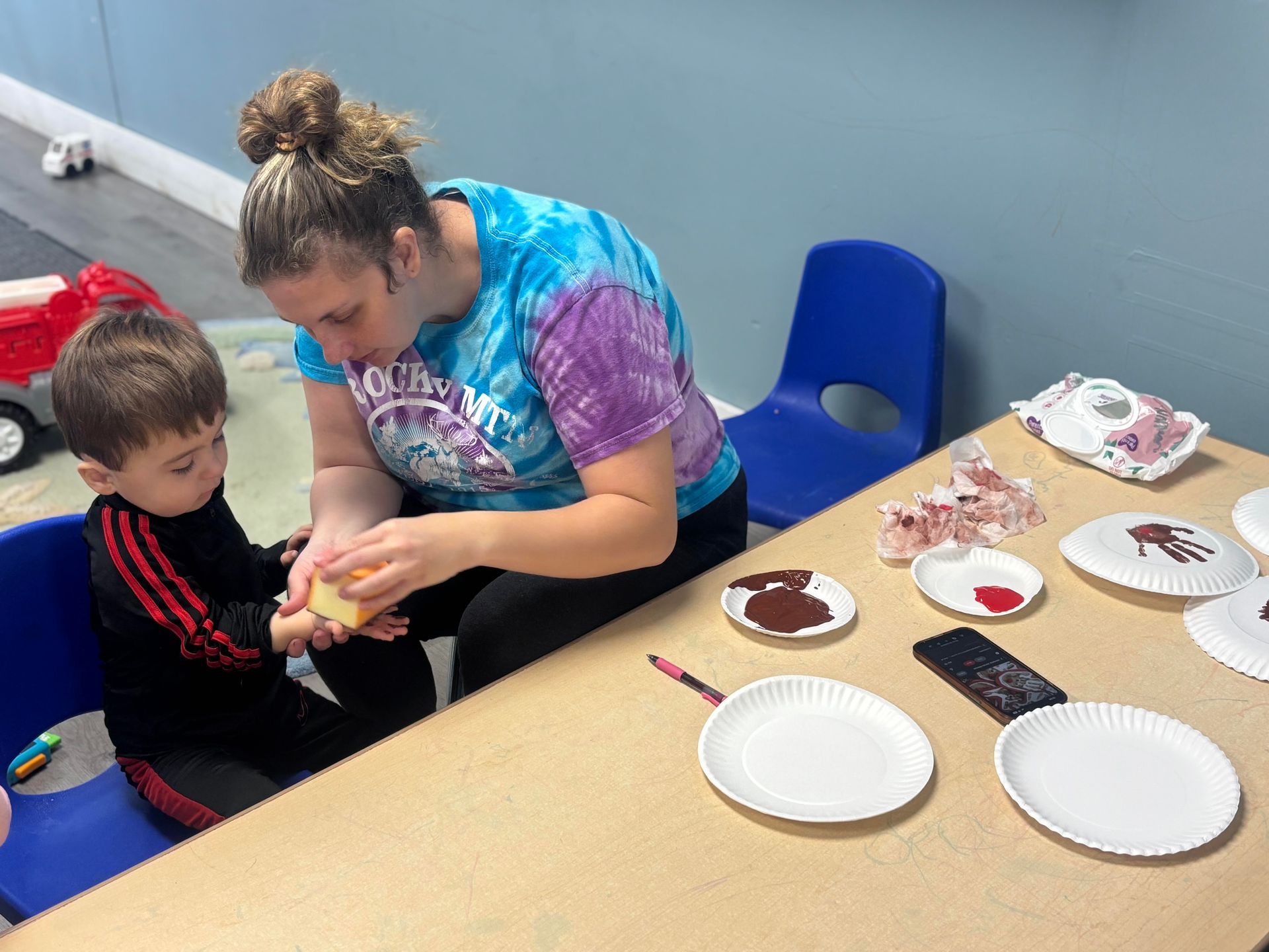 Adult and child making handprint art at a table with paper plates, paint, and materials.