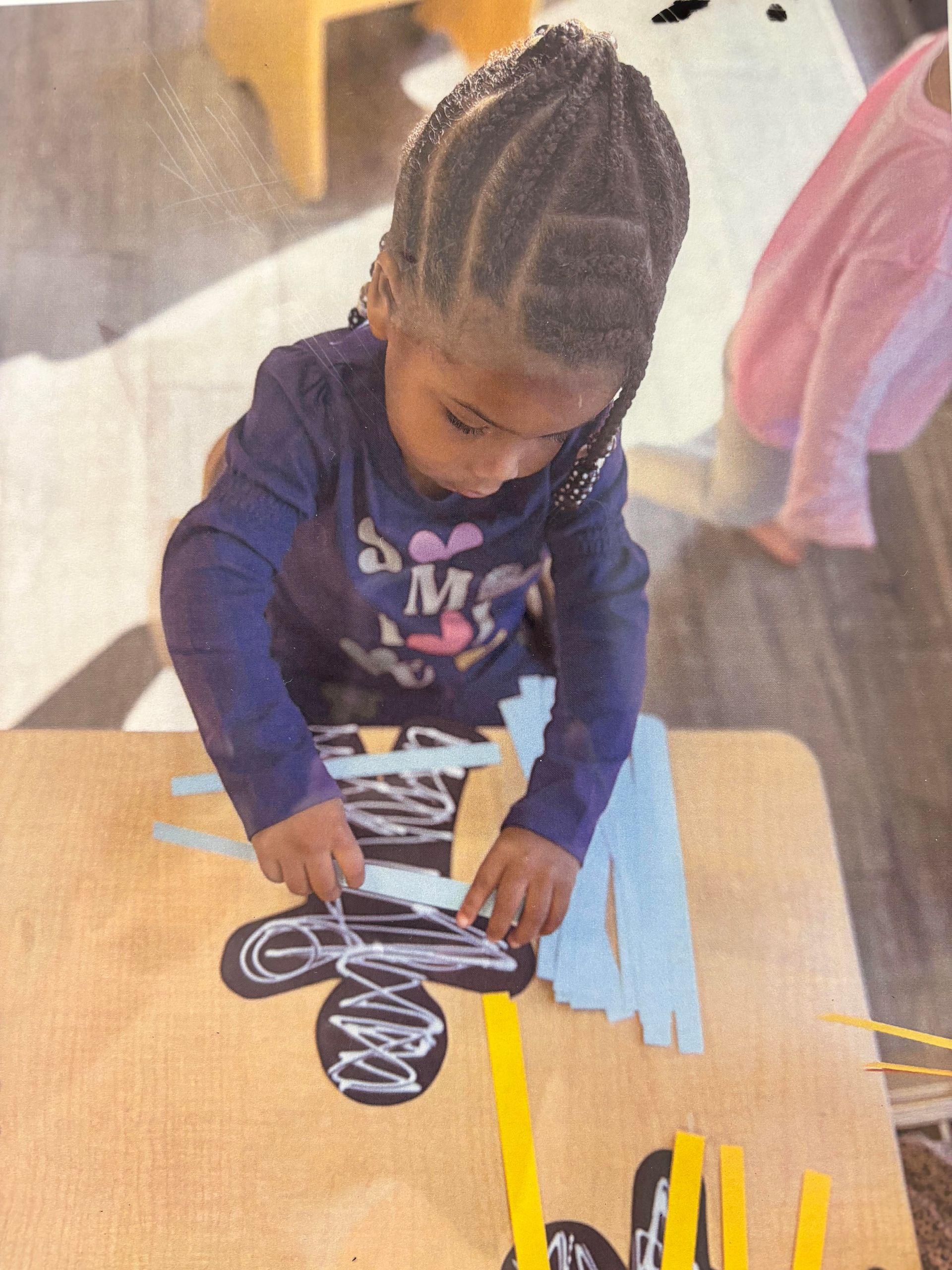 Child working on a craft at a table with paper strips and a black cutout, indoor setting.