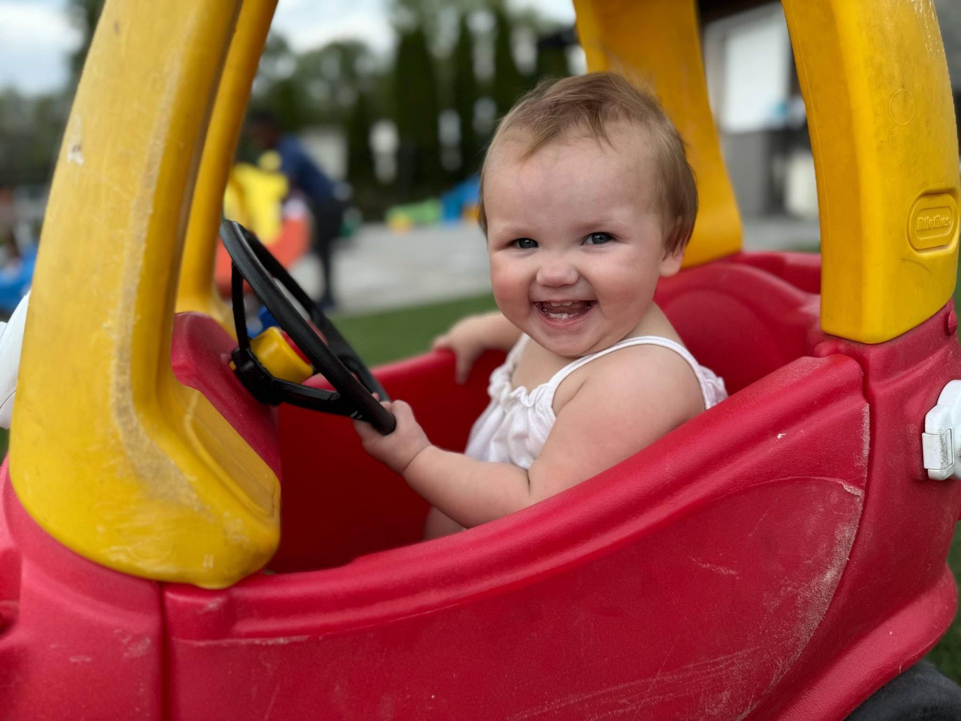 Smiling baby in a red and yellow toy car, holding the steering wheel outdoors.