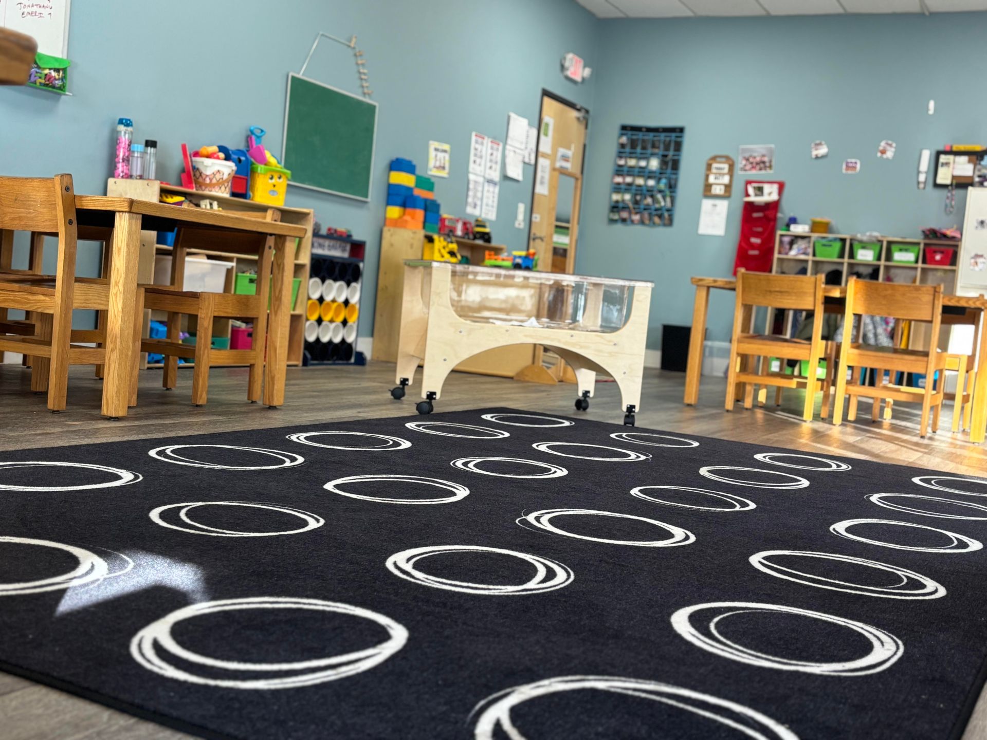 A brightly lit classroom with tables, chairs, toys, and a black rug with white circles.