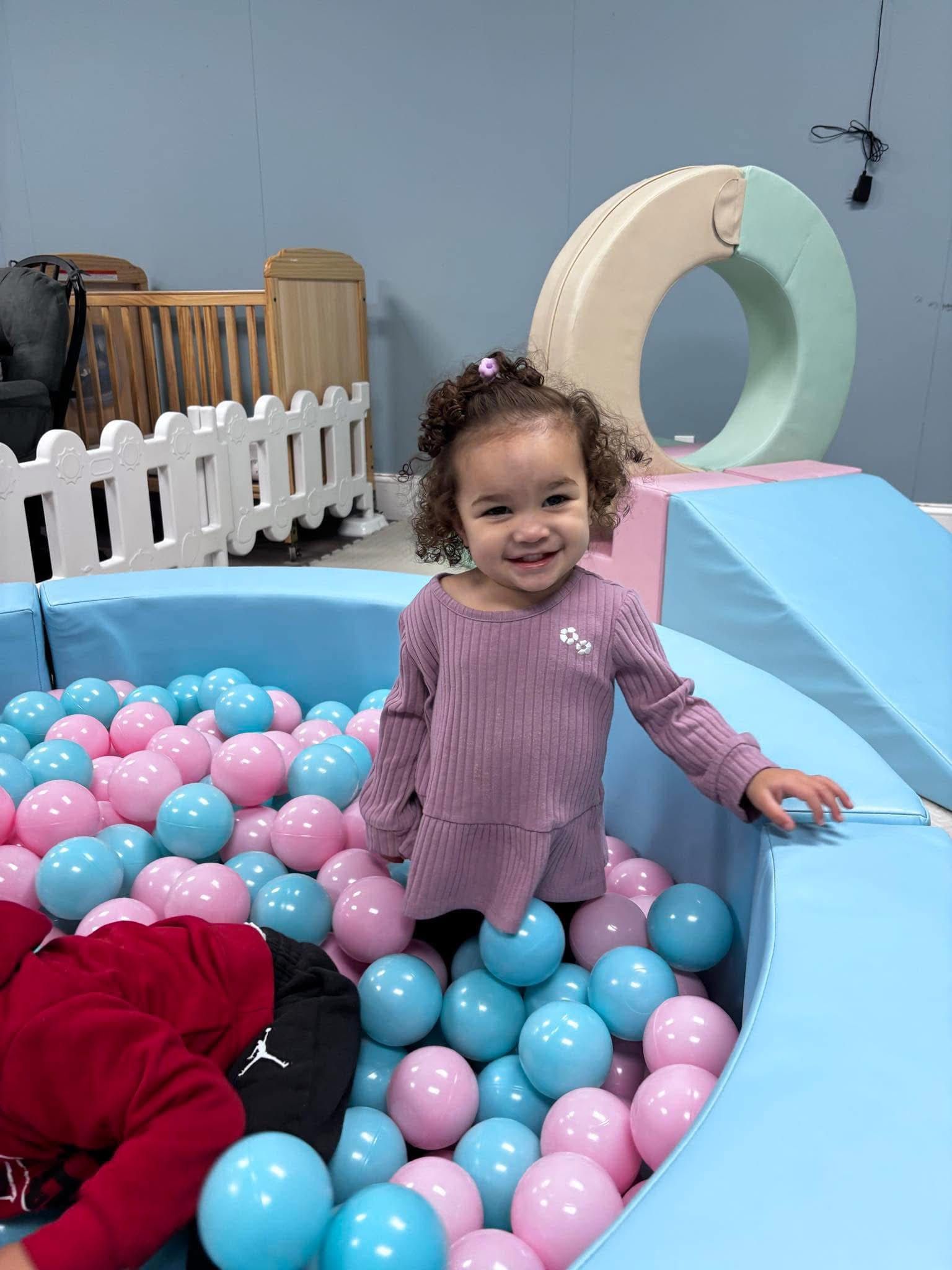 Girl smiling in a blue ball pit filled with pink and blue balls.