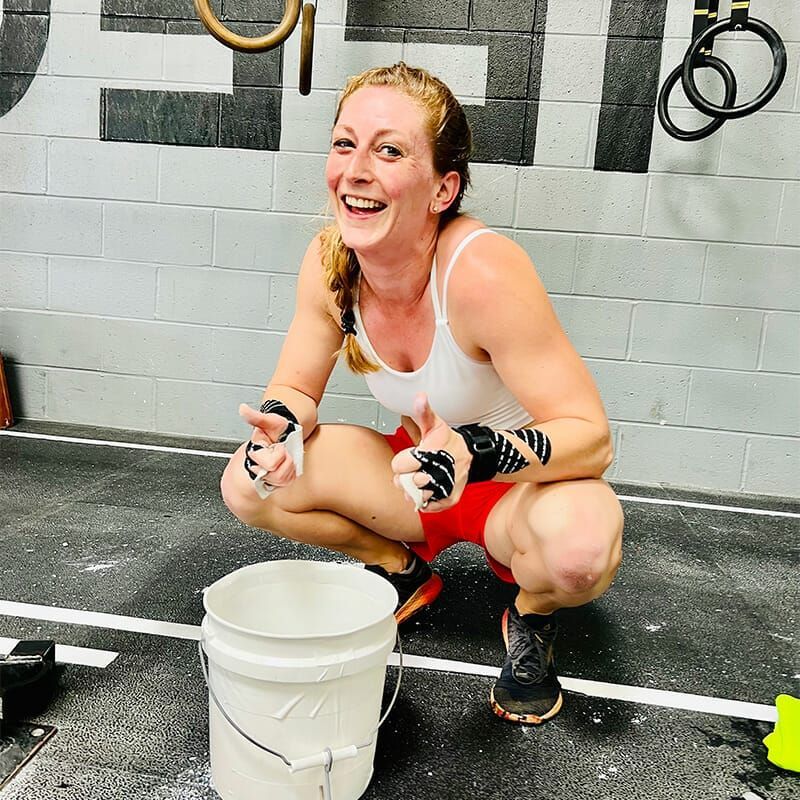 A woman is squatting down next to a white bucket and smiling.
