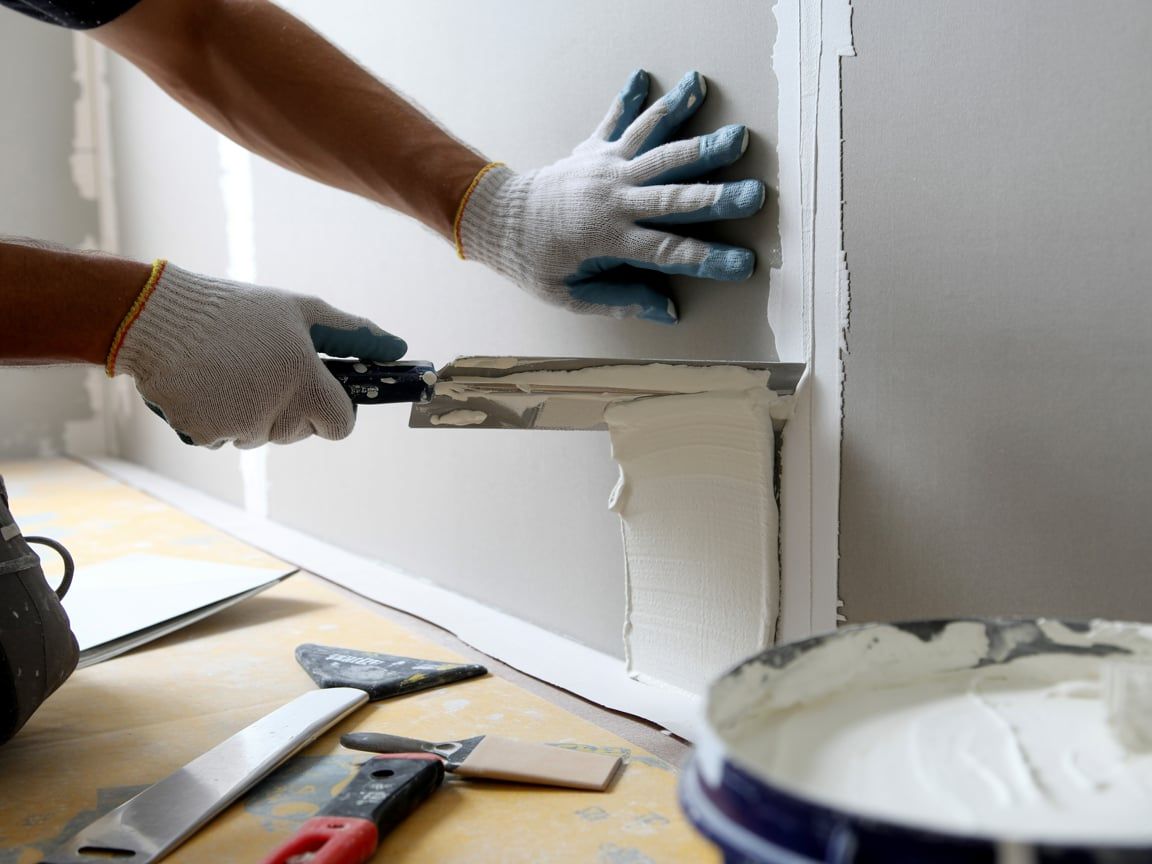 Person using a trowel to apply drywall mud on a wall, with tools and a bucket in the foreground.
