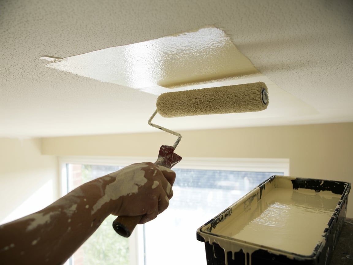 Person painting a white ceiling with a paint roller; paint tray in foreground.