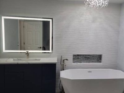A modern bathroom features a dark vanity, a backlit mirror, a freestanding white tub, and a wavy textured feature wall.