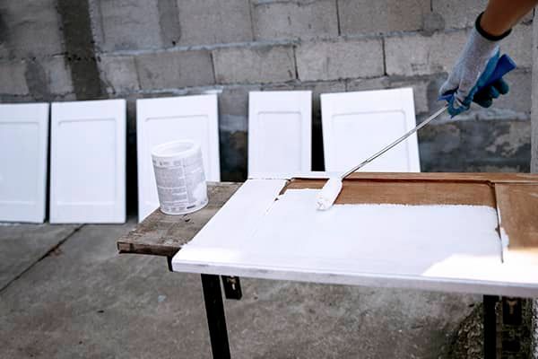 A person wearing a glove uses a foam roller to paint a wooden cabinet door white on a workbench with others drying nearby.