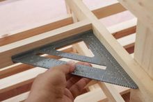 A hand uses a metal rafter square to check the 90-degree angle of a wooden frame construction.