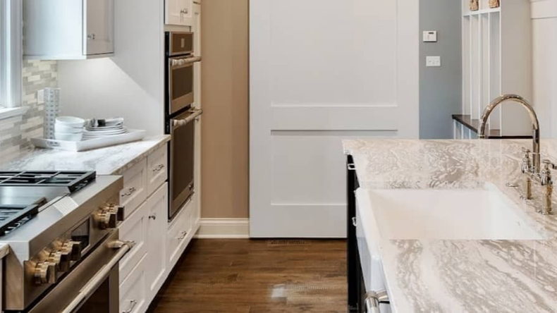 Modern kitchen featuring white cabinets, a stainless steel stove, double ovens, and a marble-topped island with a sink.