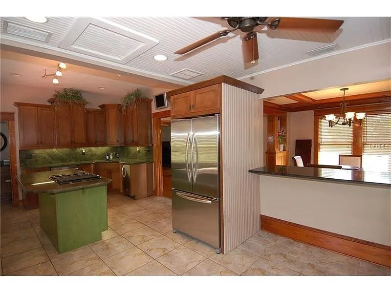 A kitchen with wood cabinetry, a green island, a stainless steel refrigerator, and a tiled floor with a ceiling fan above.
