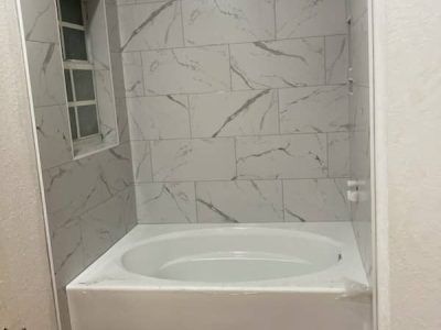 A white soaking bathtub installed in an alcove with light-colored, marble-patterned subway tile walls.
