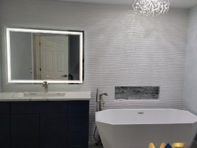 A modern bathroom with a dark blue vanity, a rectangular backlit mirror, a white soaking tub, and textured wall tiles.