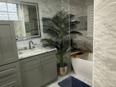 A modern bathroom with grey cabinets, a white vanity top, a large potted palm plant, and a corner soaking tub.