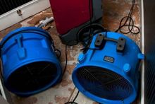 Two blue industrial floor fans and a red dehumidifier sit on a floor surface, likely used for water damage restoration.