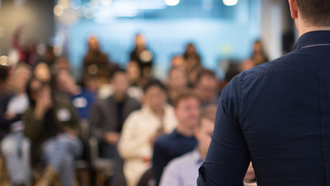 Person in blue shirt speaking to a blurry audience in a brightly lit room.