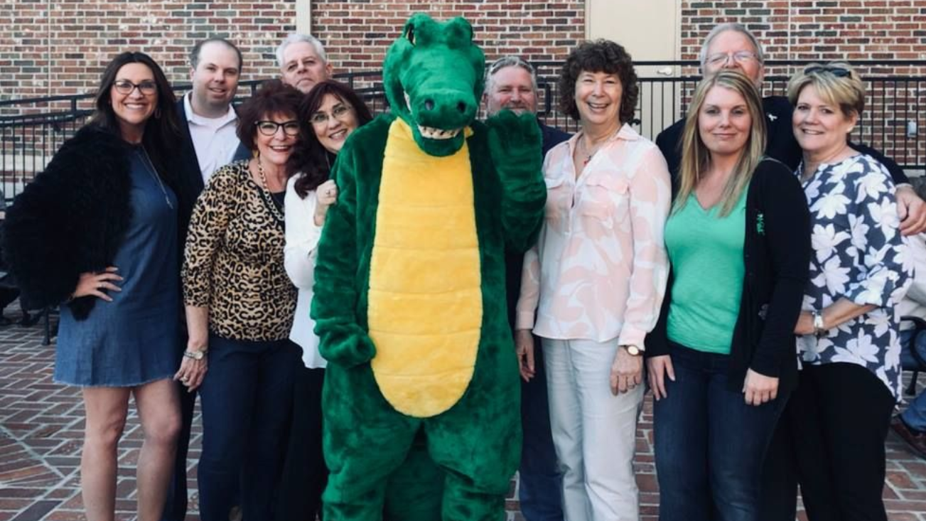 Group of people smiling with a green alligator mascot in front of a brick building.