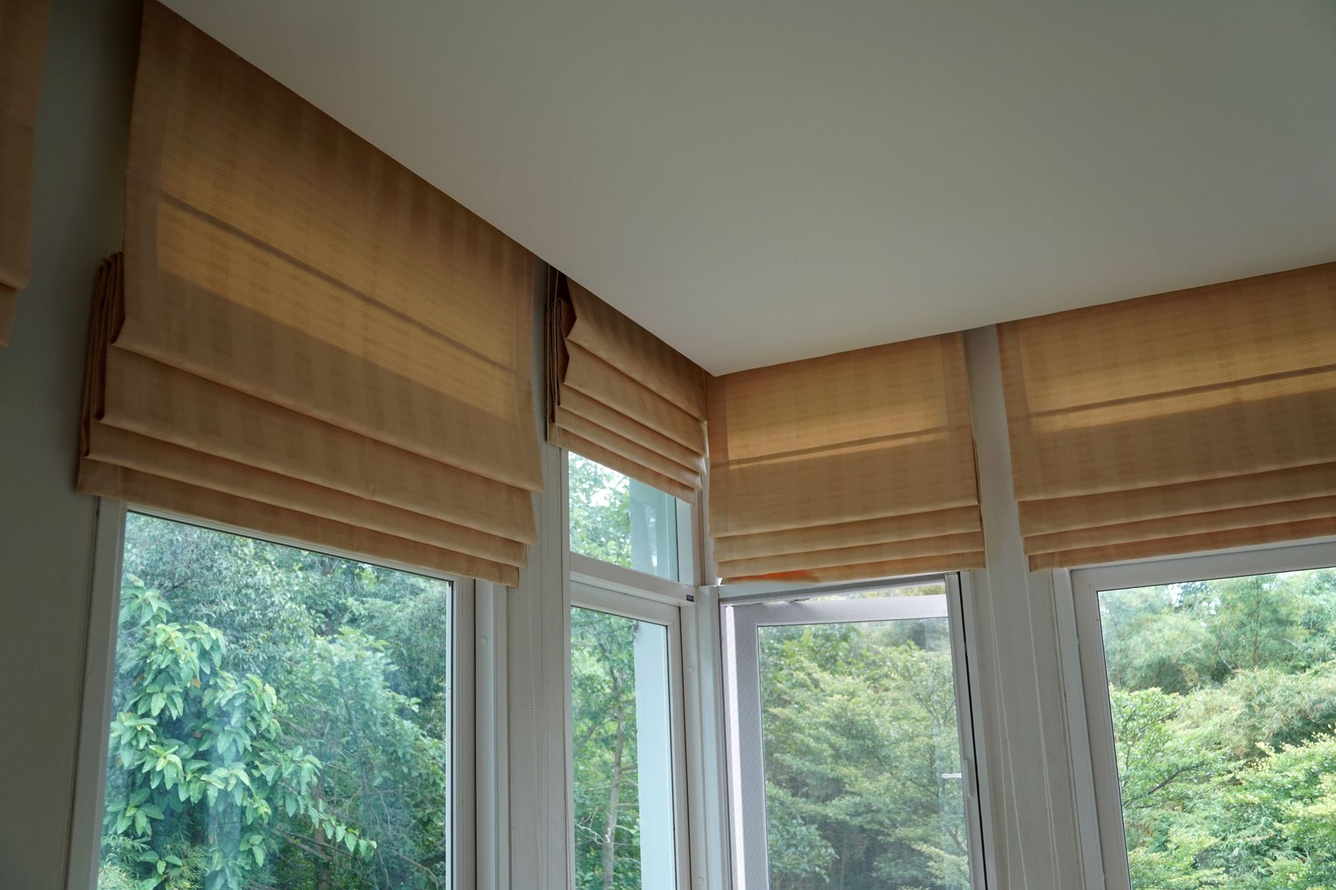 Brown Roman shades covering tall corner windows with a view of green trees.]