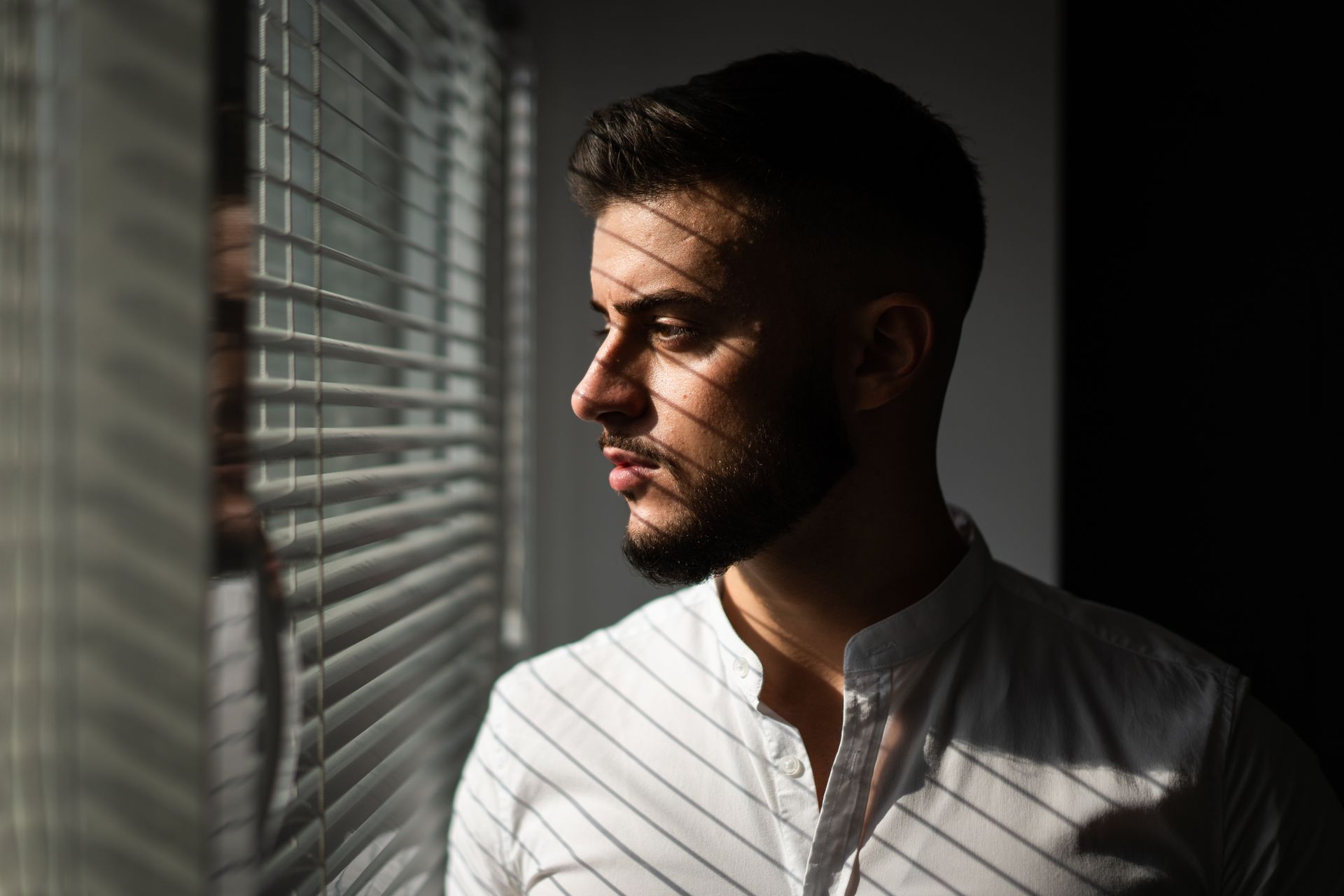 A man looking outside through slats of window shades, enjoying light and privacy.
